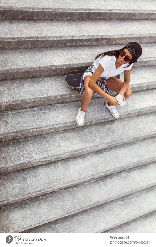 Junge Frau mit Skateboard sitzt auf einer Treppe und schaut sich um Handy Mobiltelefon Handies Handys Mobiltelefone weiblich Frauen Rollbretter Skateboards