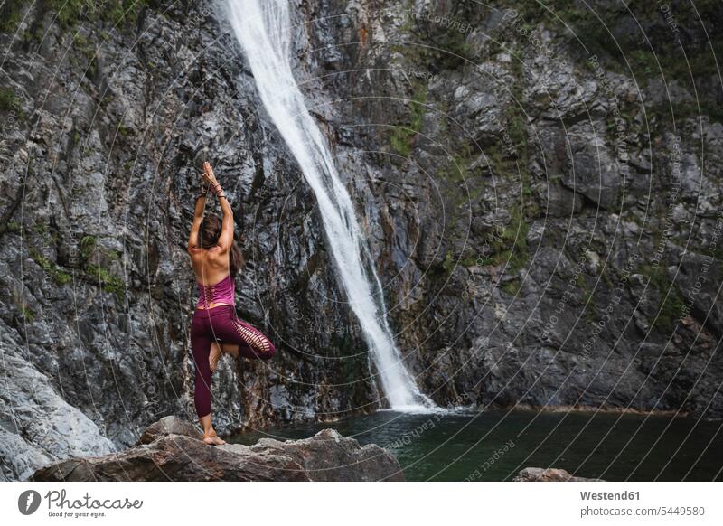 Italien, Lecco, Frau macht Baum-Yoga-Pose auf einem Felsen in der Nähe eines Wasserfalls Wasserfälle Wasserfaelle weiblich Frauen Yoga-Übungen Yogauebungen