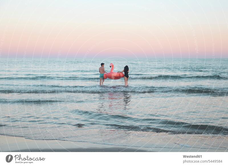 Rückenansicht eines jungen Paares, das mit einem aufblasbaren rosa Flamingo ins Meer geht Pärchen Partnerschaft Mensch Menschen Leute People Personen Meere