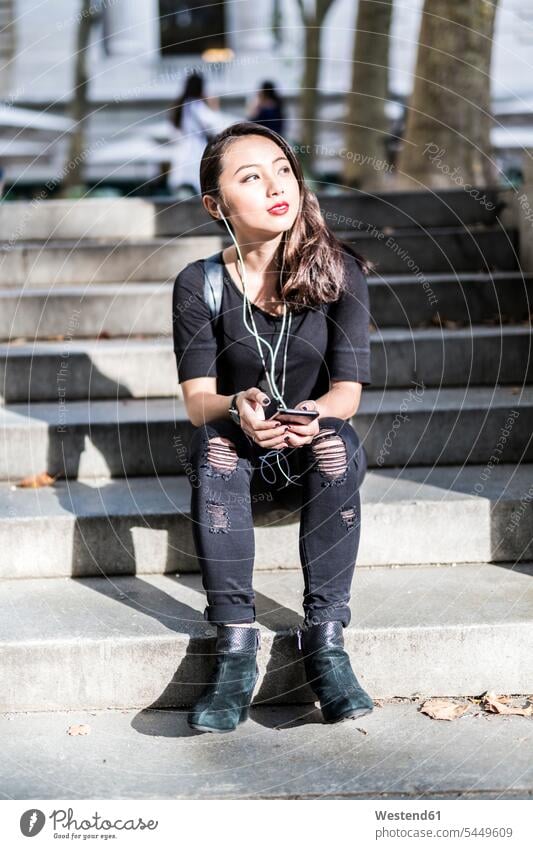 Porträt einer schwarz gekleideten jungen Frau, die auf einer Treppe sitzt und mit Kopfhörern und Handy Musik hört weiblich Frauen Erwachsener erwachsen Mensch