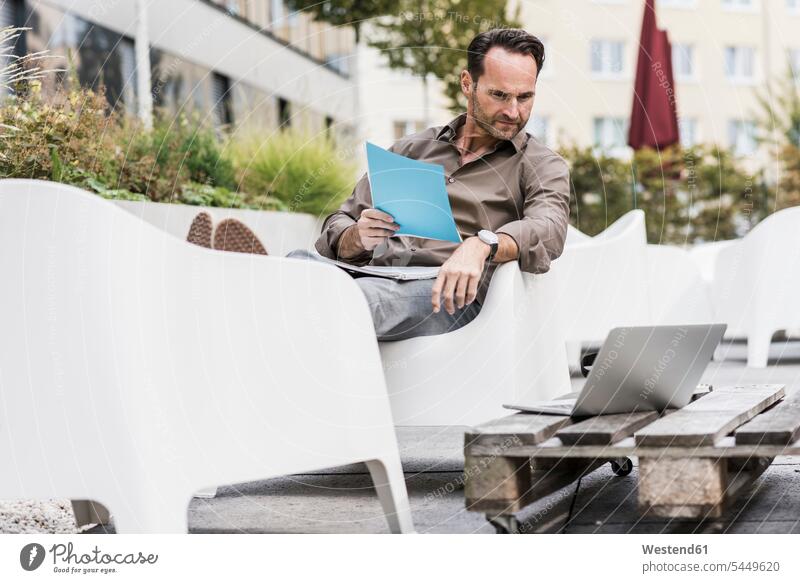 Mann mit Dokumenten sitzt auf der Terrasse und schaut auf den Laptop Geschäftsmann Businessmann Businessmänner Geschäftsmänner Notebook Laptops Notebooks