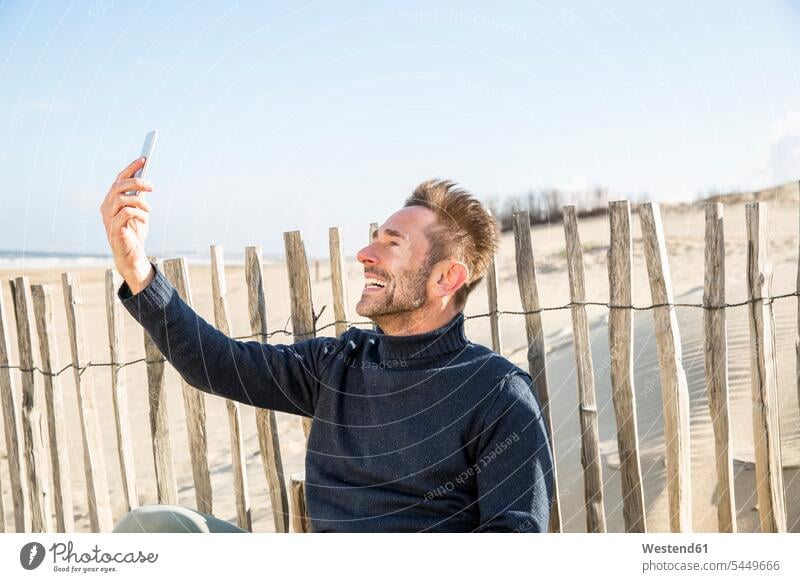 Mann macht ein Selfie am Zaun am Strand sitzen sitzend sitzt Beach Straende Strände Beaches Handy Mobiltelefon Handies Handys Mobiltelefone Selfies Männer