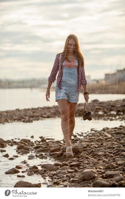 Junge Frau hält eine Kamera in der Hand und geht am steinigen Strand spazieren Fluss Fluesse Fluß Flüsse gehen gehend Fotoapparat Fotokamera lächeln weiblich