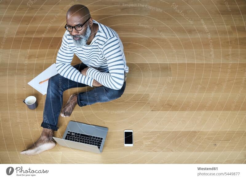 Reifer Mann sitzt auf dem Boden und arbeitet am Laptop Notebook Laptops Notebooks Smartphone iPhone Smartphones Männer männlich Zuhause zu Hause daheim barfuß