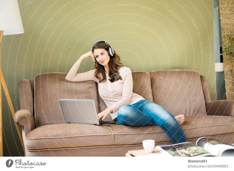 Junge Frau mit Kopfhörern sitzt mit Laptop auf der Couch Notebook Laptops Notebooks Sofa Couches Liege Sofas Kopfhoerer weiblich Frauen Computer Rechner