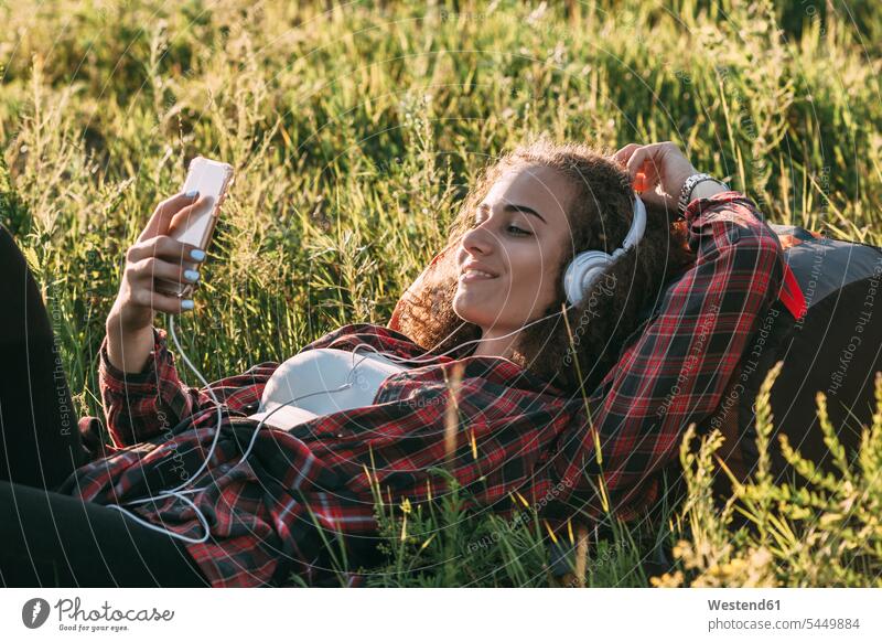 Teenagerin mit Rucksack auf einer Wiese liegend und mit Kopfhörer und Handy Musik hören junges Mädchen Teenagerinnen weiblich junge Frau Kopfhoerer Jugendliche