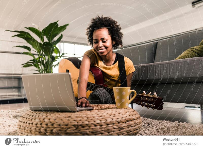 Lächelnde junge Frau zu Hause mit Gitarre und Laptop lächeln Gitarren weiblich Frauen Notebook Laptops Notebooks Saiteninstrument Saiteninstrumente