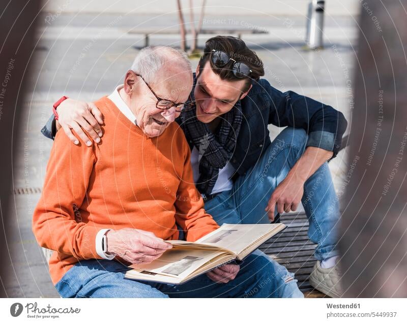 Älterer Mann und erwachsener Enkel auf einer Bank beim Betrachten eines Fotoalbums Enkelsöhne Enkelsohn sitzen sitzend sitzt Sitzbänke Bänke Sitzbank ansehen