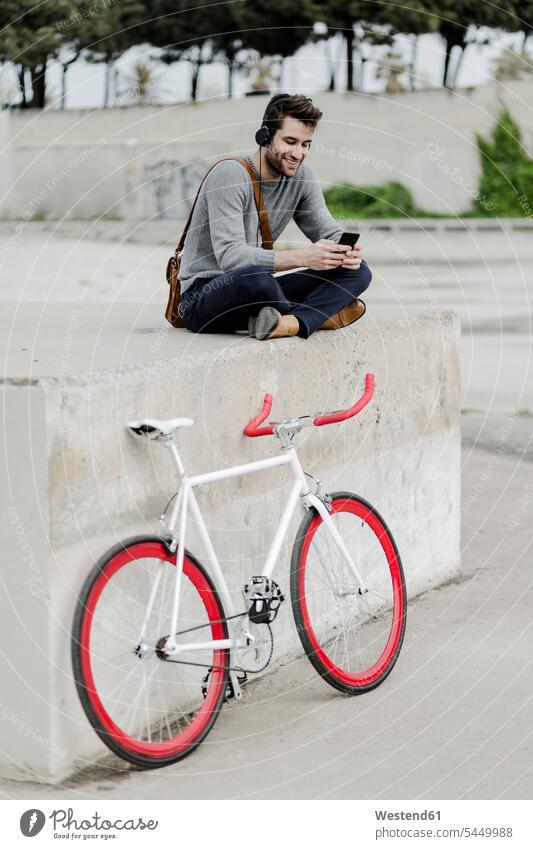 Junger Mann mit Rennrad sitzt an einer Wand und hört Musik mit Kopfhörern Männer männlich Kopfhoerer Smartphone iPhone Smartphones Erwachsener erwachsen Mensch