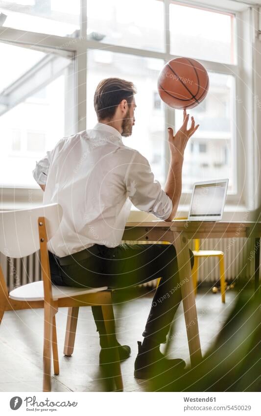 Rückenansicht eines jungen Mannes, der am Schreibtisch sitzt und auf den Laptop schaut, während er einen Basketball auf seinem Finger balanciert Notebook