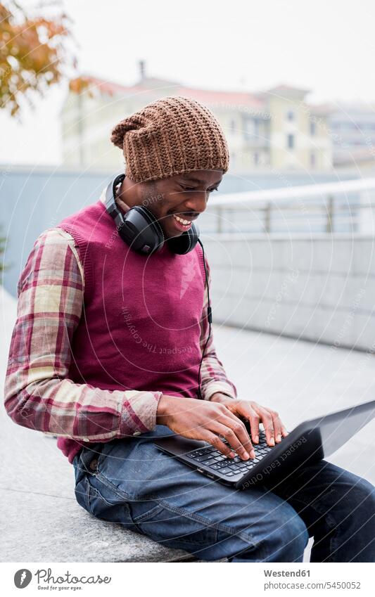 Lächelnder Mann mit Kopfhörern sitzt mit Laptop auf der Bank Männer männlich Notebook Laptops Notebooks Erwachsener erwachsen Mensch Menschen Leute People