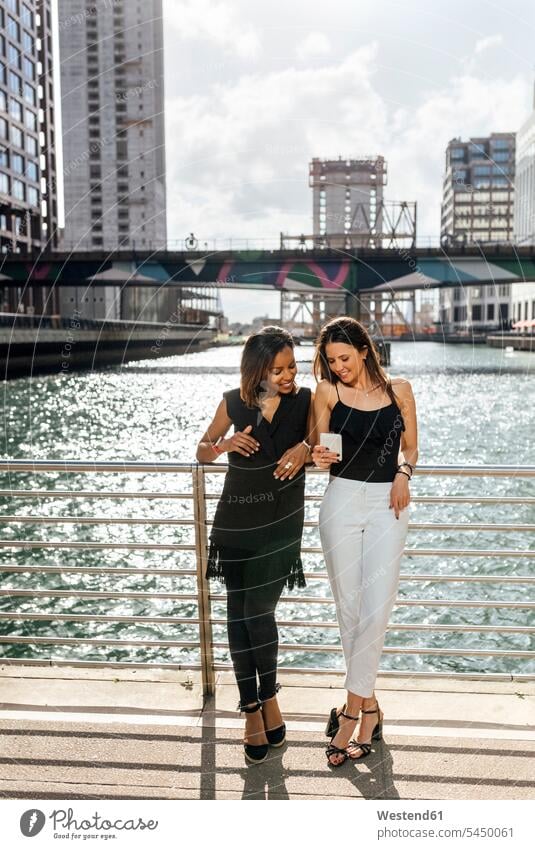 Zwei Frauen auf einer Brücke, die sich in der Stadt ein Handy teilen weiblich Kollegin Kolleginnen lächeln Mobiltelefon Handies Handys Mobiltelefone Erwachsener