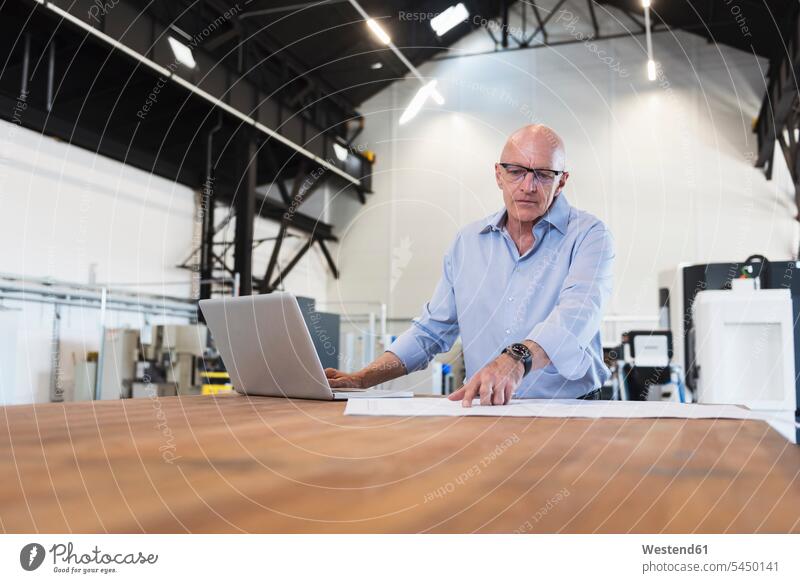 Mann mit Laptop schaut auf den Plan auf dem Tisch in der Fabrik Fabriken Notebook Laptops Notebooks Pläne Geschäftsmann Businessmann Businessmänner
