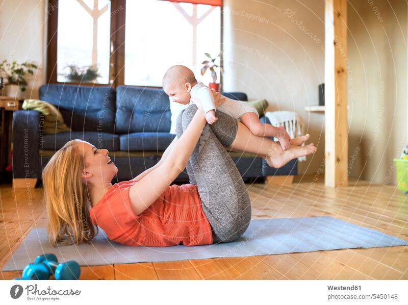 Glückliche Mutter, die ihr Baby zu Hause auf einer Matte liegend hochhebt trainieren lächeln Mami Mutti Mütter Mama heben Hantel Babies Babys Säuglinge Kind