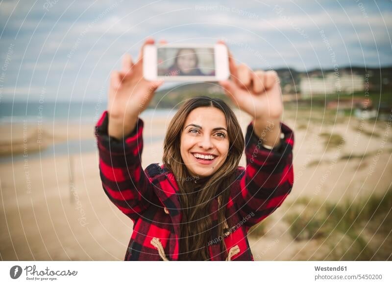 Porträt einer lächelnden Frau, die ein Selfie mit ihrem Smartphone am Strand macht Beach Straende Strände Beaches Selfies weiblich Frauen Portrait Porträts
