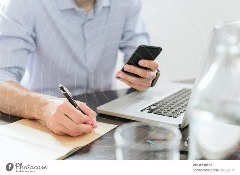 Junger Geschäftsmann, der im Büro arbeitet, mit Smartphone und Laptop Notebook Laptops Notebooks schreiben aufschreiben notieren schreibend Schrift lesen
