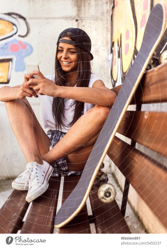 Lächelnde junge Frau mit Skateboard schaut auf Handy lächeln Rollbretter Skateboards Mobiltelefon Handies Handys Mobiltelefone Bank Sitzbänke Bänke Sitzbank