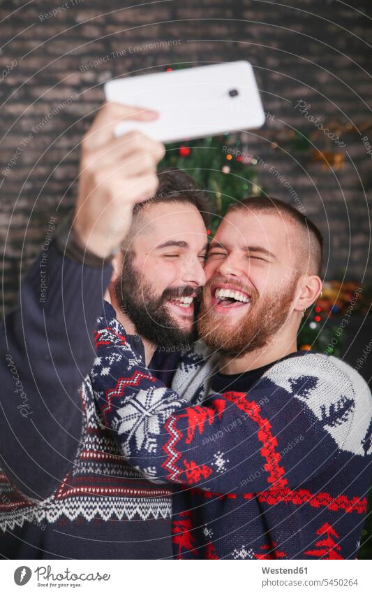 Porträt eines lachenden schwulen Paares, das zu Weihnachten zu Hause ein Selfie mit dem Smartphone macht Schwules Paar Schwuler Selfies