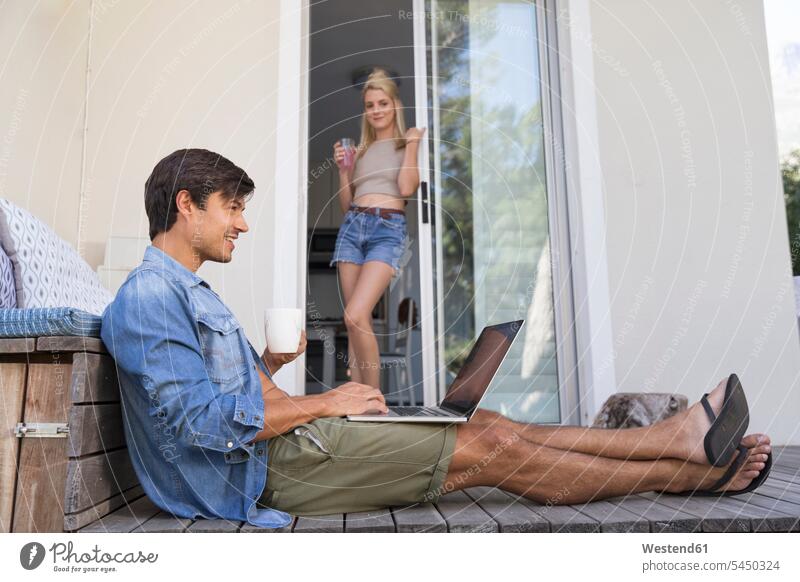 Mann sitzt mit Laptop auf der Terrasse mit Frau im Hintergrund Notebook Laptops Notebooks Männer männlich lächeln Terrassen sitzen sitzend Computer Rechner
