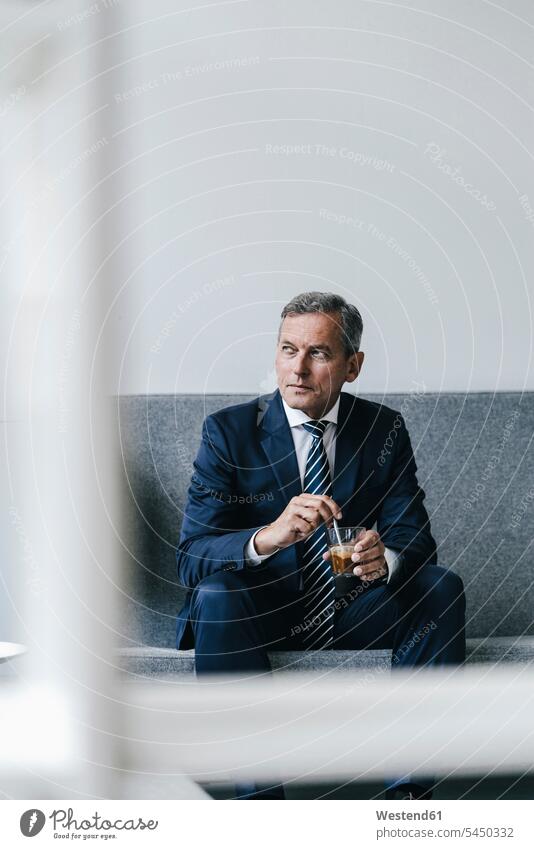 Reifer Geschäftsmann mit einem Glas Kaffee, der in seinem Büro auf der Couch sitzt und aus dem Fenster schaut Portrait Porträts Portraits Businessmann