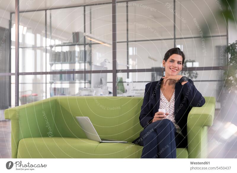 Geschäftsfrau mit Laptop, die auf einer Couch sitzt und im Büro Kaffee trinkt Smartwatch arbeiten Arbeit Internet Laptop benutzen Laptop benützen Unabhängigkeit
