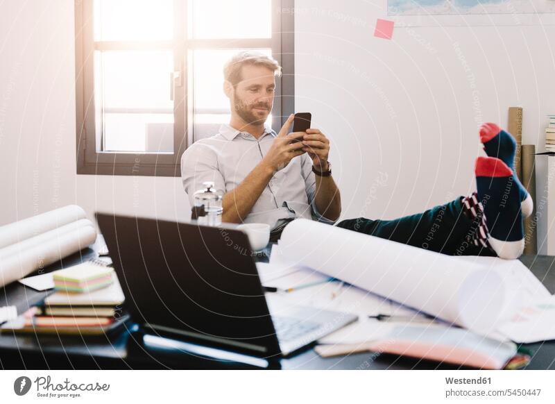 Porträt eines lächelnden Architekten, der mit erhobenen Füßen am Schreibtisch sitzt und sein Handy benutzt Büro Office Büros Arbeitsplatz Arbeitsstätte