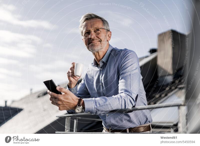 Grauhaariger Geschäftsmann mit Smartphone steht auf Balkon und trinkt Kaffee lächeln Businessmann Businessmänner Geschäftsmänner Handy Mobiltelefon Handies