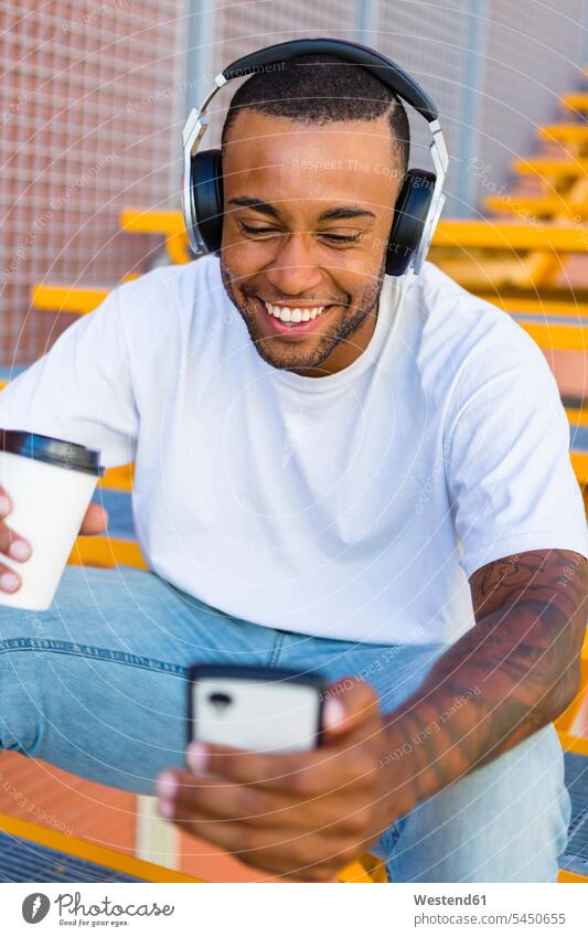 Lachender junger Mann mit Kopfhörern und Kaffee, der auf der Treppe sitzt und sich ein Smartphone ansieht Männer männlich iPhone Smartphones Kopfhoerer