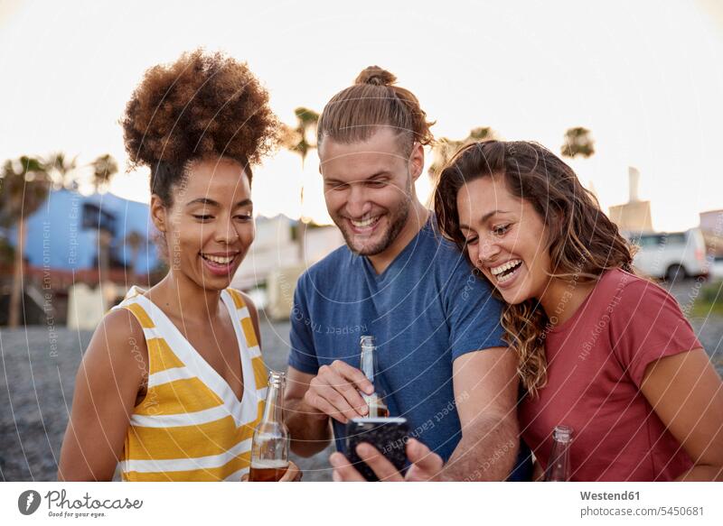 Drei lachende Freunde mit Bierflaschen beim Blick auf das Smartphone am Strand Beach Straende Strände Beaches Selfie Selfies Freundschaft Kameradschaft iPhone
