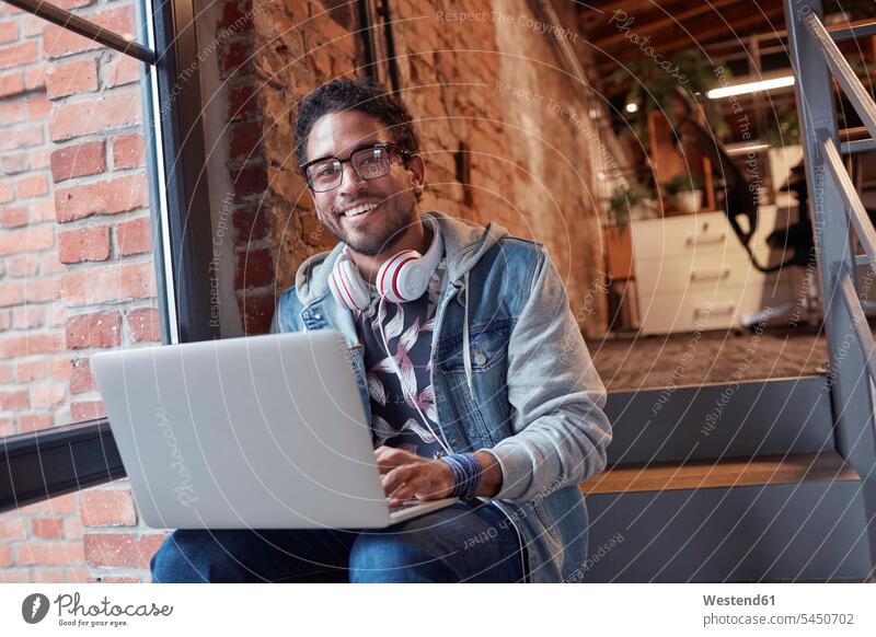 Junger Mann mit Kopfhörern sitzt auf einer Bürotreppe und benutzt einen Laptop junger Mann junge Männer Start-up Startups Start ups Start-ups Kreative Kreativer
