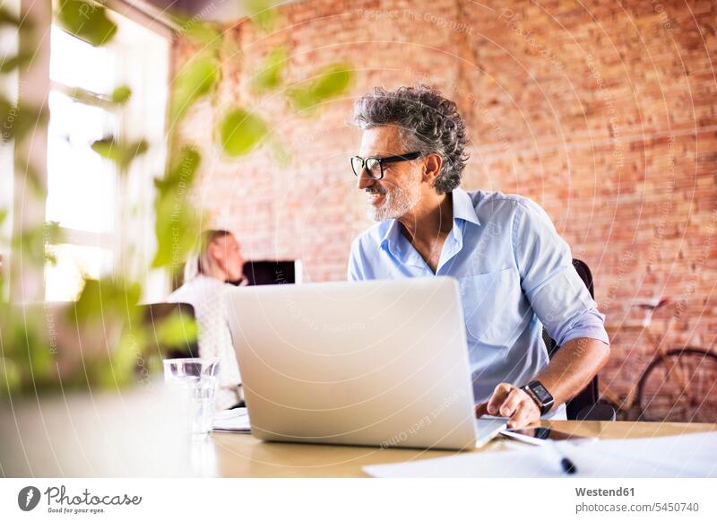 Geschäftsmann mit Laptop im Büro mit einem Kollegen im Hintergrund Schreibtisch Arbeitstisch Schreibtische Office Büros lächeln arbeiten Notebook Laptops