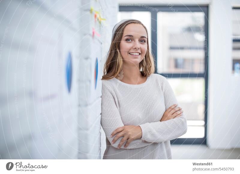 Junge Frau lehnt im Büro mit warmem Kreuz an Wand Wände Waende Büroangestellte lächeln Office Büros Zuversicht Zuversichtlich Selbstvertrauen selbstbewusst