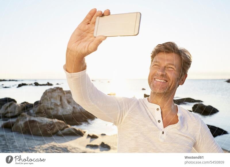 Porträt eines lächelnden Mannes, der mit seinem Handy ein Selfie am Strand macht Meer Meere Männer männlich Selfies Gewässer Wasser Erwachsener erwachsen Mensch