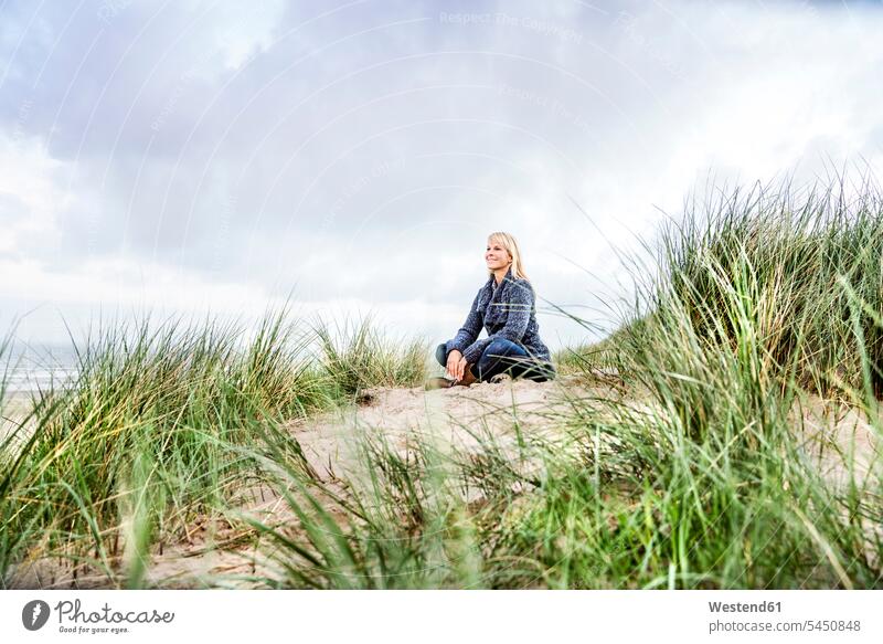 Lächelnde Frau sitzt in den Dünen Sanddüne Sanddünen sitzen sitzend weiblich Frauen lächeln Strand Beach Straende Strände Beaches Duenen Erwachsener erwachsen