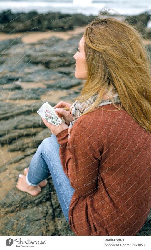 Frau mit Notizbuch sitzt im Winter auf Felsen am Strand weiblich Frauen Notizbücher Notizbuecher sitzen sitzend Beach Straende Strände Beaches Erwachsener