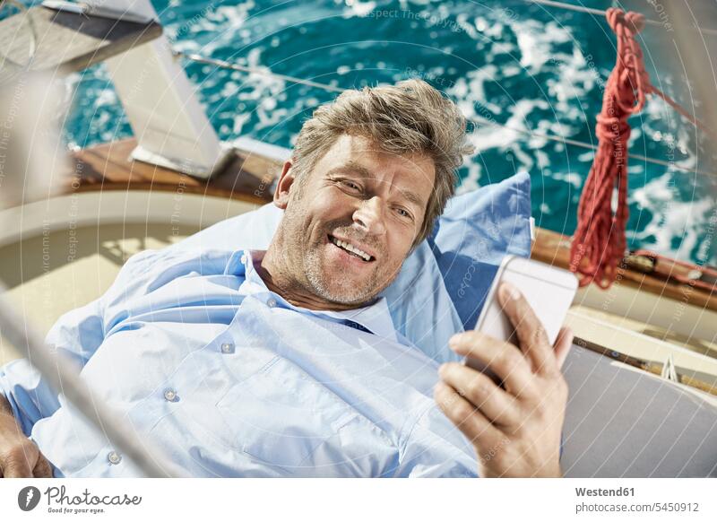 Porträt eines lächelnden reifen Mannes, der an Deck seines Segelbootes liegt und auf sein Handy schaut Männer männlich Portrait Porträts Portraits Smartphone