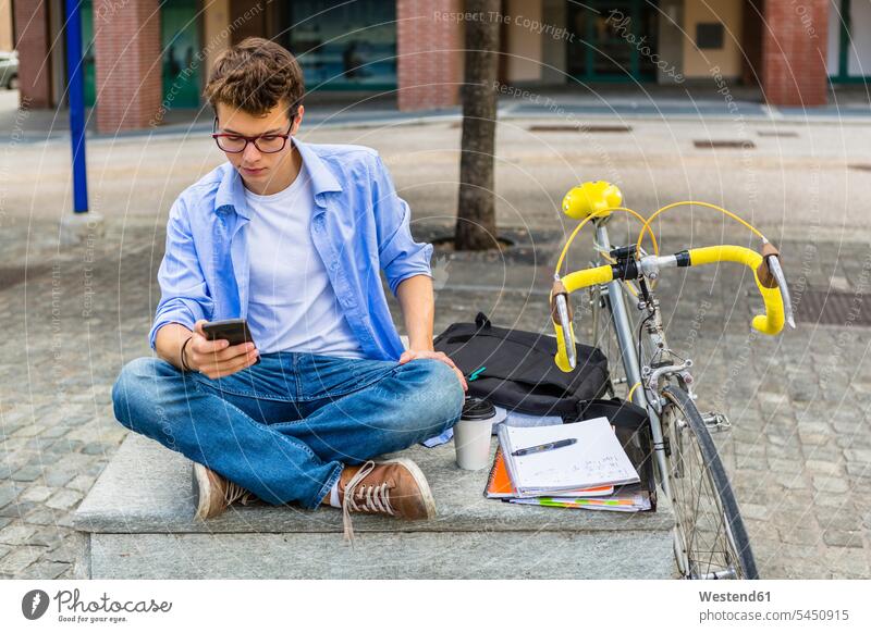 Junger Mann mit Rennrad sitzt auf Bank und schaut auf Handy Student Hochschueler Studierender Hochschüler Studenten lesen Lektüre Fahrrad Bikes Fahrräder Räder