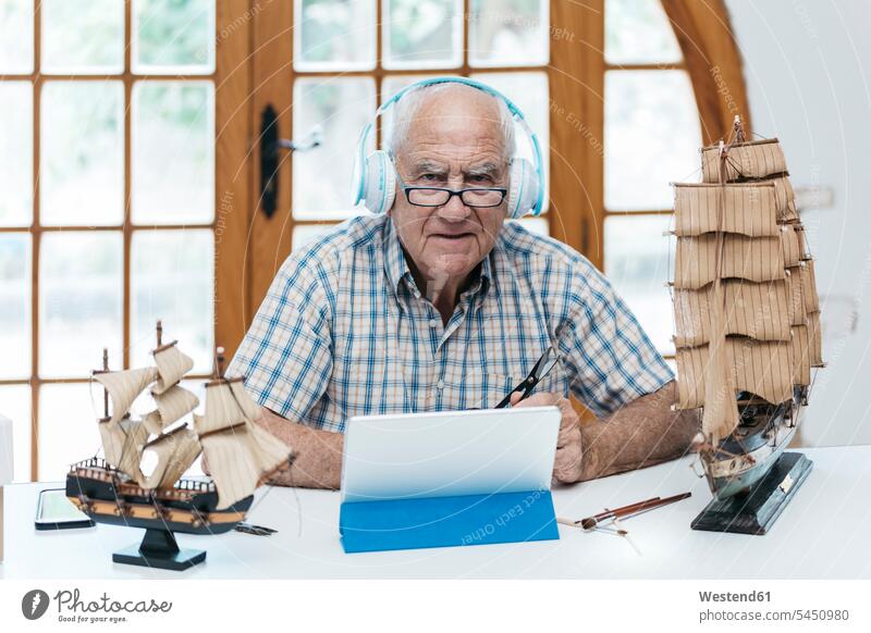 Porträt eines selbstbewussten älteren Mannes mit Kopfhörern mit Tablett und Schiffsmodell auf dem Tisch Modellboot Kopfhoerer Tablet Computer Tablet-PC
