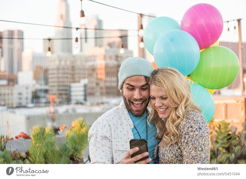 Junges Paar mit Luftballons steht auf der Dachterrasse und benutzt ein Smartphone betrachten betrachtend iPhone Smartphones Ballons Luftballone Pärchen Paare