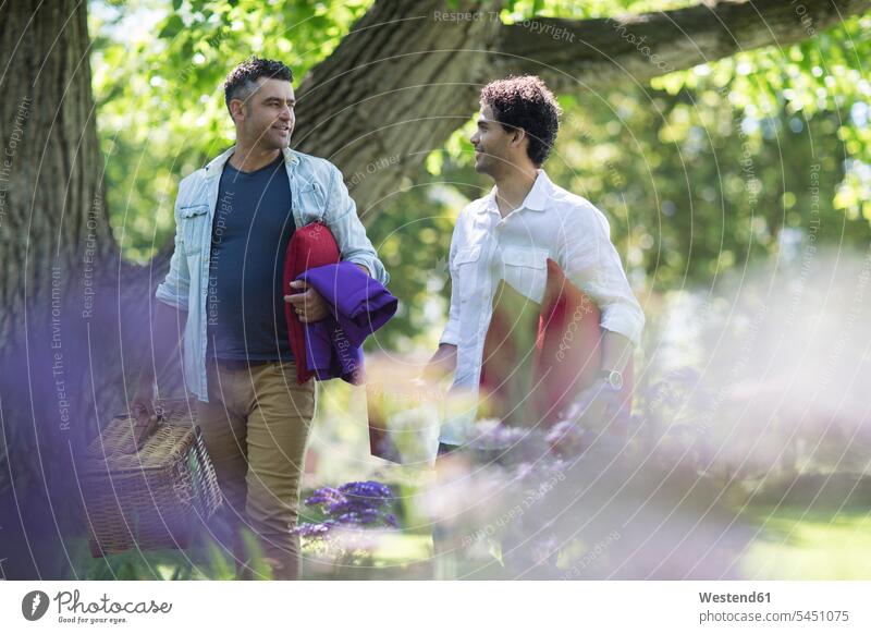 Zwei Freunde spazieren im Park mit Picknickkorb und Kissen gehen gehend geht Mann Männer männlich lächeln Erwachsener erwachsen Mensch Menschen Leute People