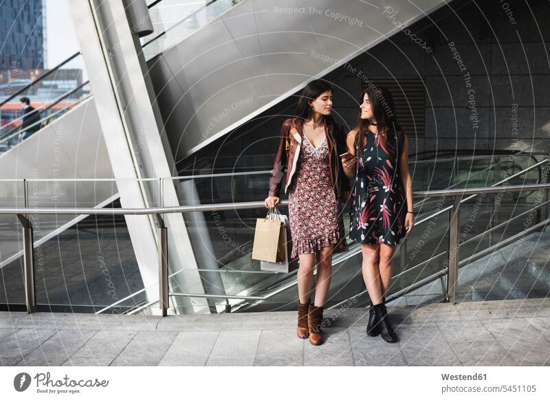 Zwei junge Frauen mit Einkaufstaschen und Handy in der Stadt lächeln Mobiltelefon Handies Handys Mobiltelefone Freundinnen Telefon telefonieren Kommunikation