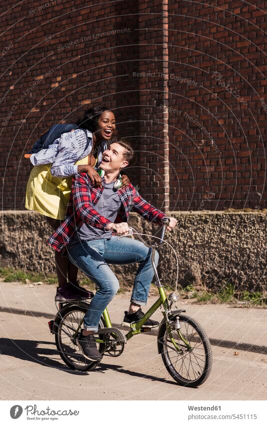 Junger Mann fährt Fahrrad mit seiner Freundin auf dem Gepäckträger stehend aktiv radfahren fahrradfahren radeln jung multikulturell Gemeinsam Zusammen