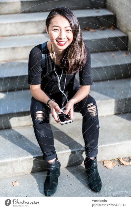 Porträt einer schwarz gekleideten jungen Frau, die auf einer Treppe sitzt und mit Kopfhörern und Handy Musik hört lächeln weiblich Frauen Erwachsener erwachsen