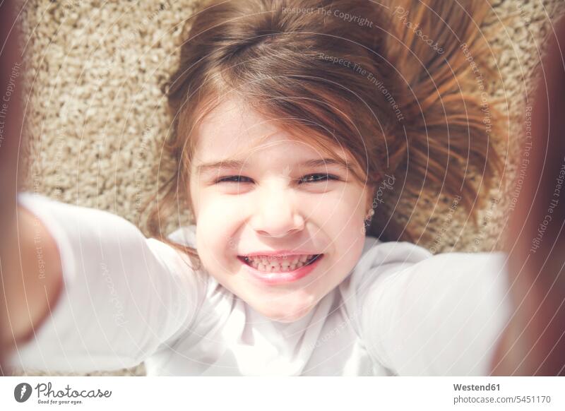 Porträt eines grinsenden kleinen Mädchens, das auf dem Teppich liegt und ein Selfie macht weiblich Teppiche Portrait Porträts Portraits Kind Kinder Kids Mensch