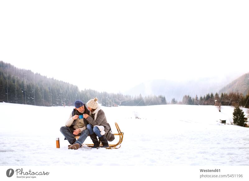 Älteres Ehepaar sitzt nebeneinander auf Schlitten in schneebedeckter Landschaft Seniorenpaar älteres Paar Seniorenpaare ältere Paare Seniorenpärchen Schnee