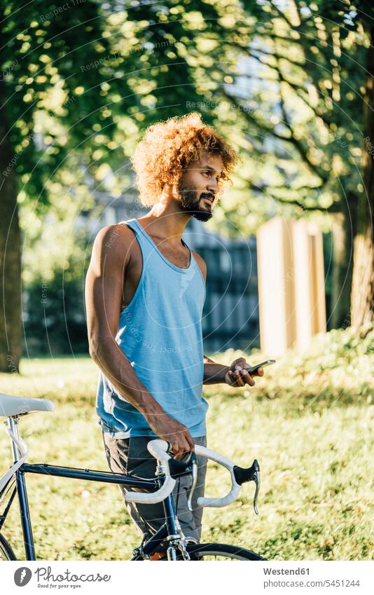 Junger Mann mit Fahrrad und Handy im Park Bikes Fahrräder Räder Rad Männer männlich Mobiltelefon Handies Handys Mobiltelefone Raeder Erwachsener erwachsen