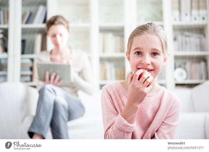 Mädchen isst Apfel zu Hause mit der Mutter im Hintergrund weiblich lächeln essen essend Familie Familien Zuhause daheim Äpfel Aepfel Kind Kinder Kids Mensch