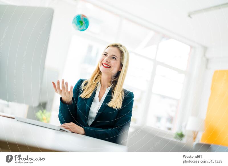 Lächelnde Geschäftsfrau am Schreibtisch, die einen Mini-Globus hochwirft lächeln Büro Office Büros Weltkugel Erdkugel Globen Geschäftsfrauen Businesswomen