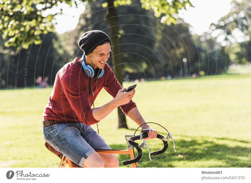 Lachender Mann auf Rennrad mit Blick auf Handy in einem Park Fahrrad Bikes Fahrräder Räder Rad Parkanlagen Parks Männer männlich Raeder Erwachsener erwachsen