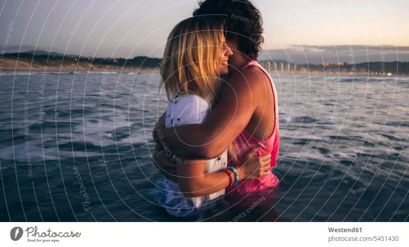 Junges Paar umarmt sich in der Abenddämmerung im Meer Meere lächeln umarmen Umarmung Umarmungen Arm umlegen Pärchen Paare Partnerschaft Gewässer Wasser Mensch